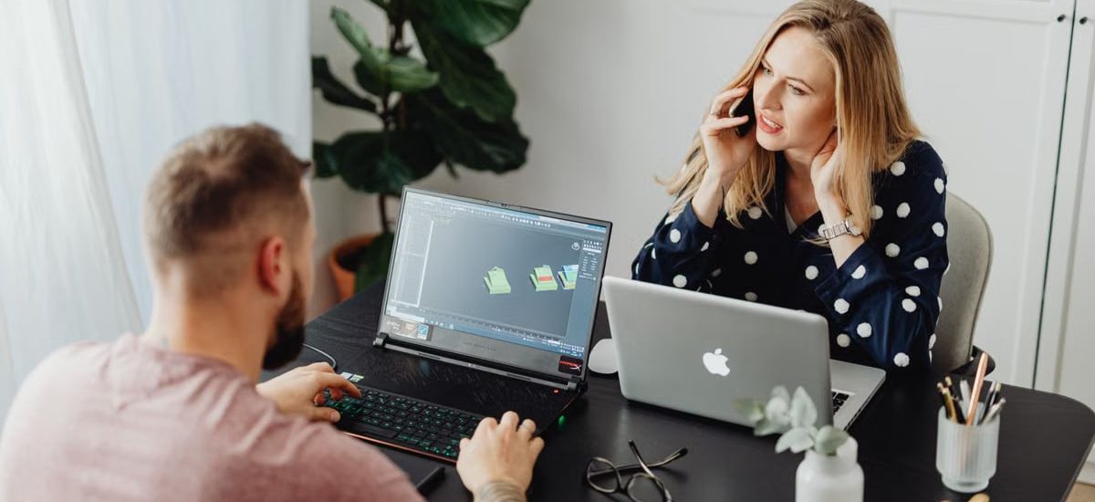 Two coworkers collaborating with laptops and phone in modern office setting.