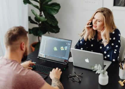 Two coworkers collaborating with laptops and phone in modern office setting.